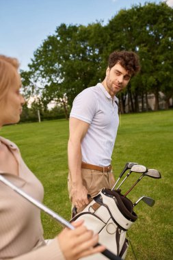 Zarif giyinmiş genç bir çift yemyeşil bir yeşil ile çevrili bir golf sahasında yan yana durur..