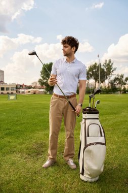 Şık giyinmiş bir adam golf sahasında, açık gökyüzünün altında, yemyeşil bir çantayla duruyor..