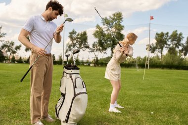 Zarif giyinmiş bir erkek ve kadın yemyeşil bir alanda golf oynuyorlar. Zarafet ve beceri sergiliyorlar..