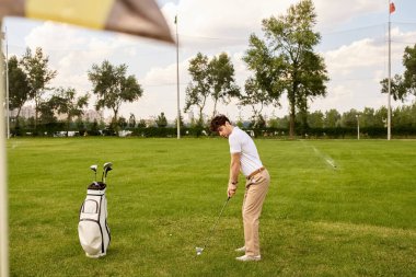 Zarif giyimli bir adam yemyeşil bir tarlada golf topuna vuruyor, eski para tarzında zarafet ve incelik sergiliyor..