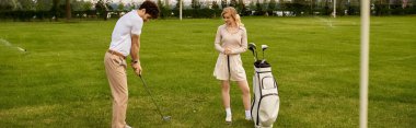 Zarif giyinmiş genç bir çift yemyeşil bir alanda golf oynuyor. Birlikte güzel bir ortamda vakit geçiriyorlar..