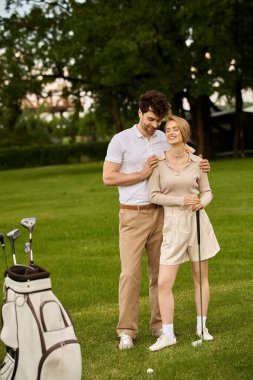 Zarif giyinmiş şık genç bir çift yemyeşil bir golf sahasında yan yana durur..