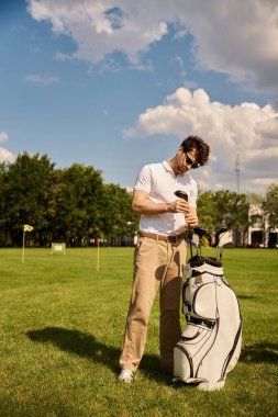 Zarif giyinmiş bir adam lüks bir golf kulübündeki yemyeşil bir alanda golf çantasının yanında zarif bir şekilde duruyor..