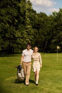 Zarif, genç bir çift şık giyinmiş, yemyeşil bir yeşil alanla çevrili bir golf sahasında sakin bir yürüyüş yapıyor..