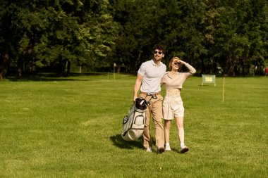 Tarz sahibi genç bir çift, resim gibi bir golf sahasında rahatça yürüyüş yapar, birbirlerine eşlik ederek eğlenirler..