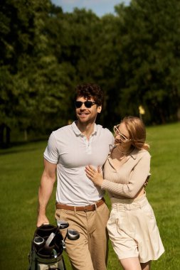 A stylish young couple in elegant attire leisurely walking with a golf bag in a lush park setting. clipart