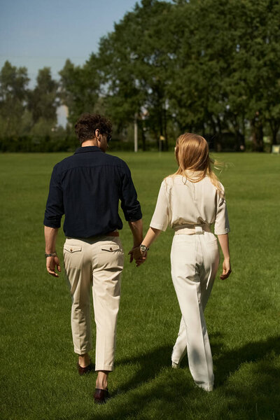 A young couple in elegant attire holding hands while walking through a lush park together.