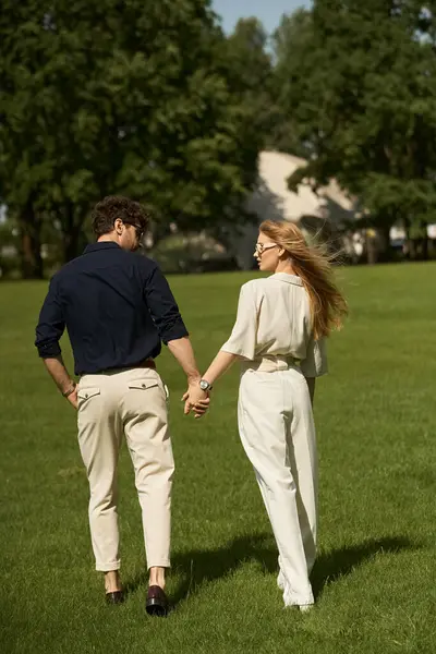 stock image A beautiful young couple in elegant attire, holding hands while standing in a lush green field.