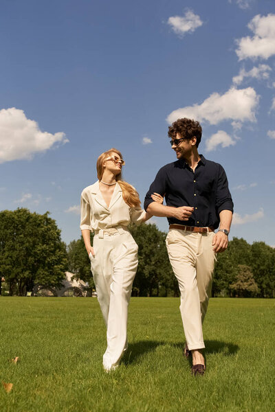 A beautiful young couple in elegant clothing stroll through a green field, embodying a luxurious lifestyle.