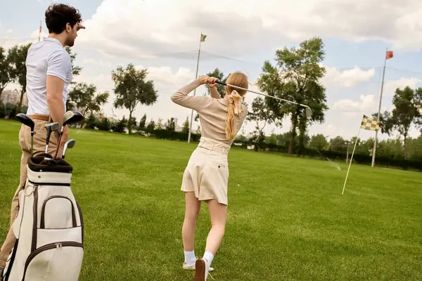 Zarif giyinmiş bir erkek ve kadın, saygın bir golf kulübünün huzuruyla çevrili yemyeşil bir alanda golf oynuyorlar..