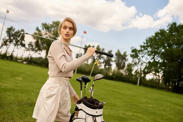 Şık giyinmiş genç bir kadın, üst sınıf bir golf kulübündeki yemyeşil bir alanda golf çantası tutuyor..