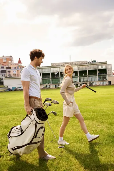 stock image A man and woman in elegant attire walking casually on a grassy field with golf clubs in hand.