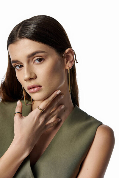 A beautiful, stylish woman with long dark hair poses in a stunning green dress and gold earrings against a gray backdrop.