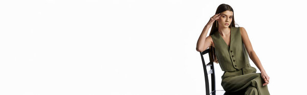 Модная женщина с длинными темными волосами, сидящая в кресле, рука об лицо, глубоко погруженная в мысли на сером фоне.