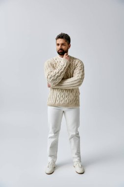 Sakallı bir adam beyaz bir süveter ve pantolonla gri bir stüdyo arkaplanına karşı sofistike bir şekilde yayılır..