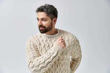 Sakallı karizmatik bir adam gri bir stüdyo arkaplanına beyaz bir süveter giyip poz veriyor..