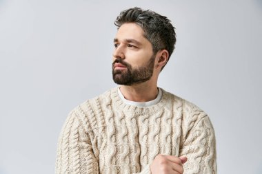 A charismatic man with a beard strikes a pose in a white sweater against a grey studio background. clipart