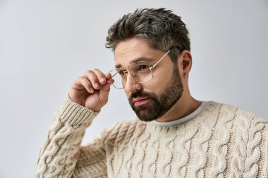 Sakallı karizmatik bir adam, beyaz bir kazakla stüdyoda gri bir arka plana karşı poz veriyor..