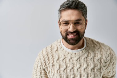 A bearded man exudes allure in a white sweater, complemented by glasses, on a grey backdrop in a studio setting. clipart