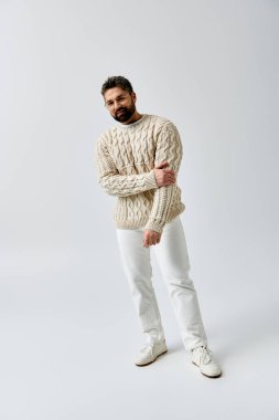 A charismatic man with a beard strikes a pose in a fashionable white sweater against a simple grey studio backdrop. clipart