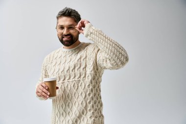 A bearded man in a white sweater delicately holds a steaming cup of coffee against a grey studio backdrop. clipart