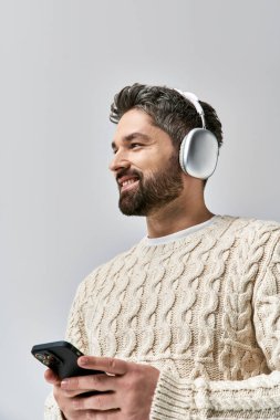 Beyaz kazaklı sakallı bir adam kulaklık takıyor. Gri bir arka plana takılı cep telefonu ekranına takılı..
