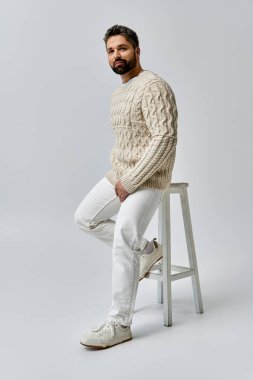 A bearded man exudes charm as he sits on a stool, elegantly clad in a white sweater against a grey studio backdrop. clipart