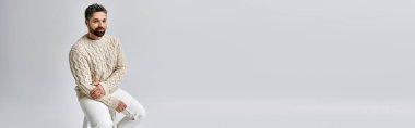 Çekici bakışları olan sakallı bir adam beyaz bir süveter ve pantolonla gri bir stüdyo arkaplanına poz veriyor..