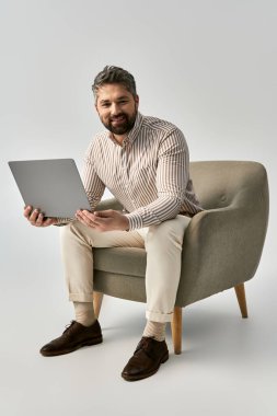 A stylish man with a beard is seated on a chair, working on his laptop in a trendy and sophisticated setting. clipart