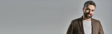 Zarif giyinmiş şık sakallı bir adam stüdyo ortamında parlak, gri bir arka planda duruyor..