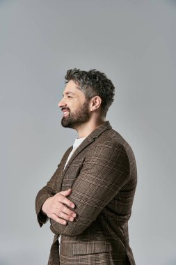 Zarif giyinmiş, sakallı sofistike bir adam, kolları gri bir arkaplanda duruyor..