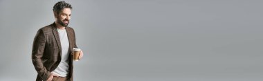 Sakallı şık bir adam zarif bir şekilde elinde bir fincan kahveyle gri bir stüdyo arkaplanına karşı duruyor..