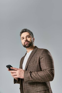 Takım elbiseli sakallı bir adam cep telefonu tutuyor. Gri bir arka planda stüdyoda zarafet yayıyor..