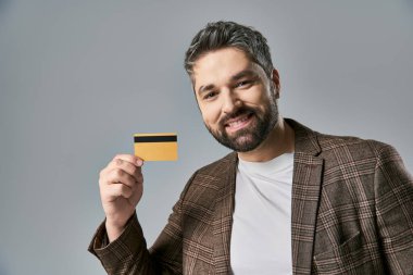 A stylish man with a beard holding a credit card and smiling joyfully against a grey studio background. clipart