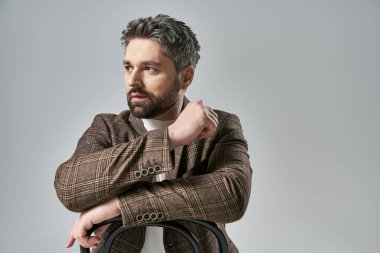 Zarif giyinmiş sakallı bir adam, kollarını kavuşturmuş, gri bir stüdyonun arka planına yaslanmış..