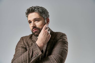 Tarafsız gri bir arka plana karşı, toprak tonlu ceketli şık görünümlü, sakallı bir adam..