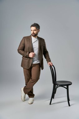 Sakallı şık bir adam stüdyo ortamında parlak siyah bir sandalyenin yanında duruyor..