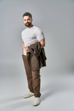 Sakallı bir adam, beyaz tişört ve kahverengi pantolonuyla gri bir stüdyo geçmişine karşı zarafet yayar..