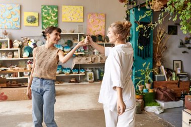 Two women, a loving and tender lesbian couple, stand in an art studio, holding hands. clipart