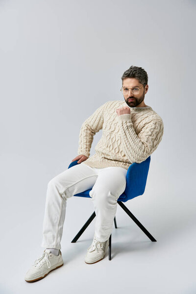 A bearded man in a white sweater sitting in a chair, deep in thought with his hand on his chin.