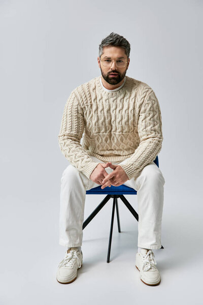 A bearded man in a white sweater striking a pose while sitting on a blue chair in a studio with a grey background.