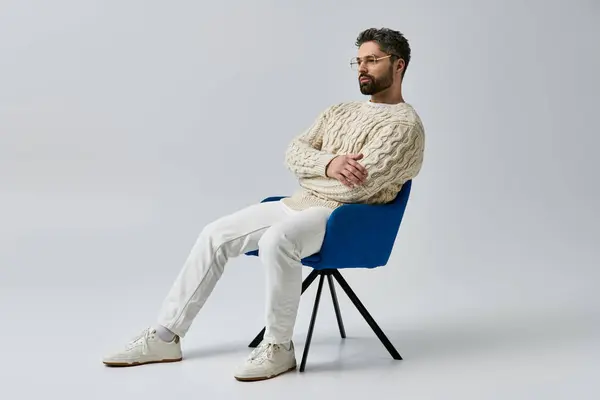 stock image A man with a beard sits in a chair, arms crossed, wearing a white sweater on a grey studio background.