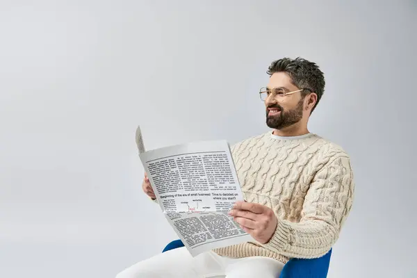 一个留着胡子的时髦男人坐在椅子上 专心致志地阅读报纸 在工作室里 他的背景是灰色的 — 图库照片