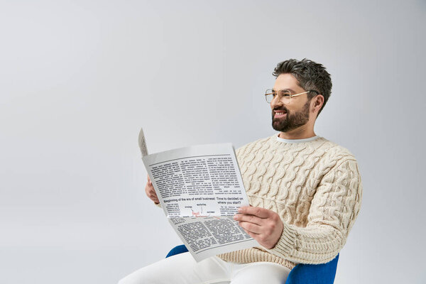 Стильный мужчина с бородой сидит на стуле, погруженный в чтение газеты, на сером фоне в студии.