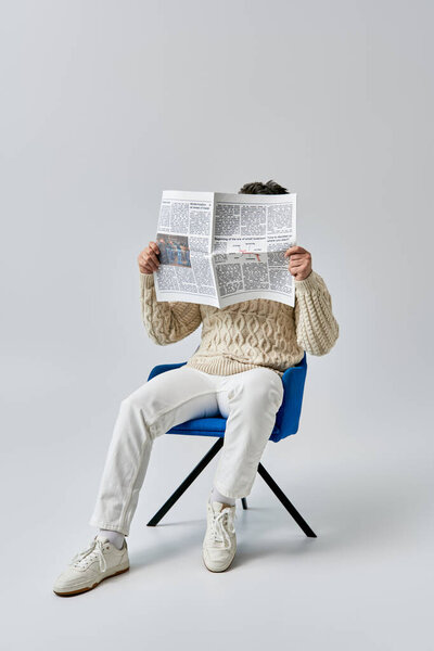 Мужчина в белом свитере, сидящий в кресле, сосредоточился на чтении газеты..