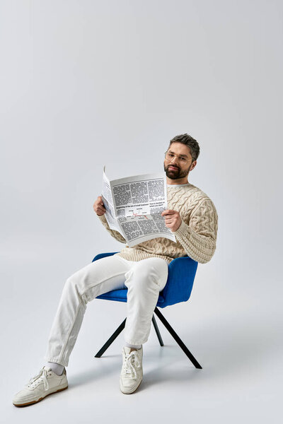 Стильный мужчина с бородой, сидящий в кресле, погруженный в чтение газеты на сером фоне.