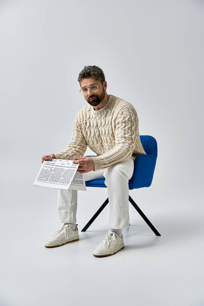 Стильный мужчина с бородой, в белом свитере, сидит в кресле и читает газету на сером фоне..