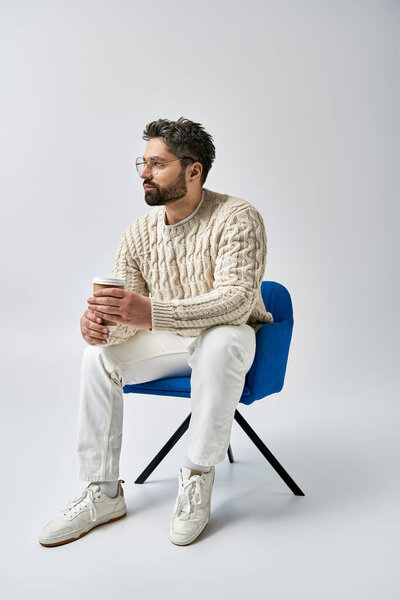 A bearded man in a white sweater sits on a blue chair, crossing his arms in a contemplative pose against a grey background.