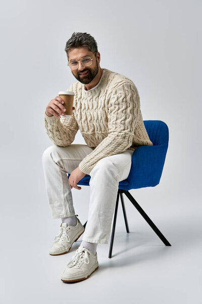 A stylish man with a beard sits on a chair in a white sweater, savoring a cup of coffee on a grey studio backdrop.
