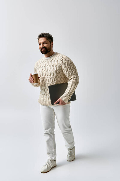 A bearded man in a white sweater holds a laptop and a cup of coffee, lost in thought.